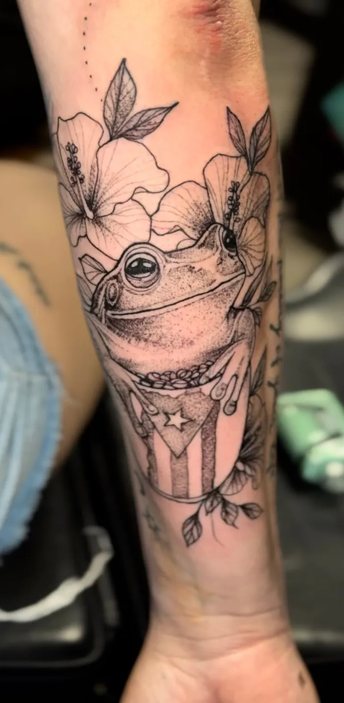 Fine line forearm tattoo of a coqui frog with hibiscus flowers and the Puerto Rico flag in black and grey by Duk Tattoo, Boca Raton, Florida